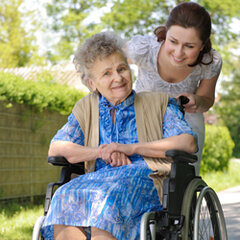 Eine ältere Dame im Rollstuhl wird durch einen Sommergarten gefahren. 