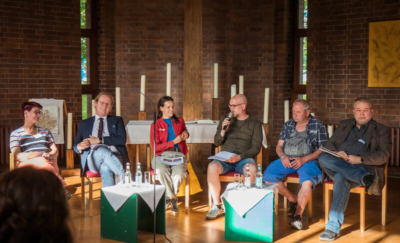 Rednerinnen und Redner sitzen auf Stühlen im Halbkreis zum Publikum gerichtet und unterhalten sich über Obdachlosigkeit in Lichtenberg