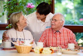 Eine ältere Frau und ein älterer Mann sitzen am Frühstückstisch. Eine Pflegerin begrüßt sie.
